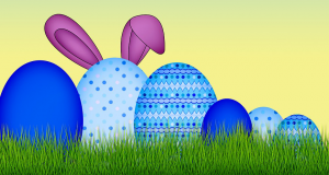 Blue decorated eggs on a bed of grass with bunny ears behind the largest egg.