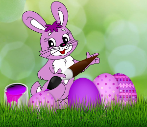 Easter Bunny holding a paint brush with purple decorated eggs on the grass.