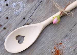 Wooden spoon with heart cut out of the middle. A small pink rose bud is tied on the handle. Spices spread around the spoon on a wooden background.