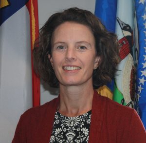 Rebecca Gay, short dark hair in a standing in front of the American and Currituck County Flags.